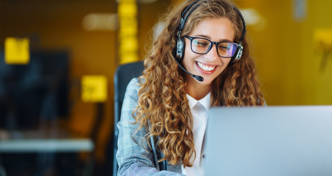 A women in business attire wearing glasses and headset smiles as she looks at a computer screen