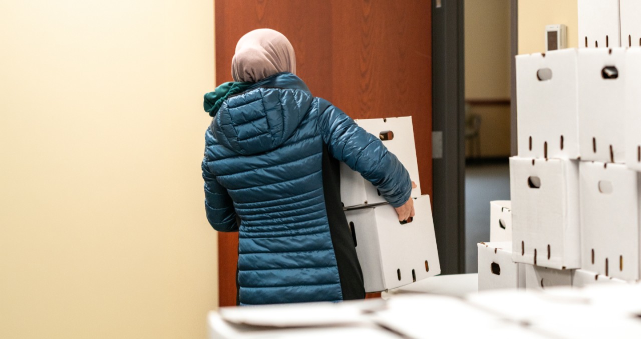 A woman in a blue coat and headscarf picks up two white boxes from a large stack of boxes