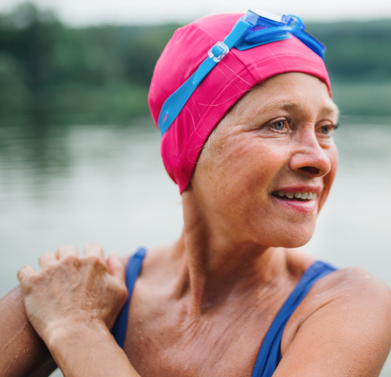 A woman wearing a pink swim cap and blue swimsuit stands and smiles with a lake in the background
