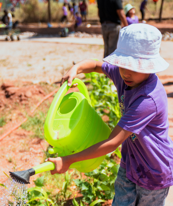 A young boy wearing a bucket hat waters plants with a large green watering can