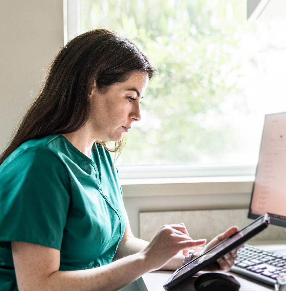 A nurse looks at a tablet while sitting at a computer desk