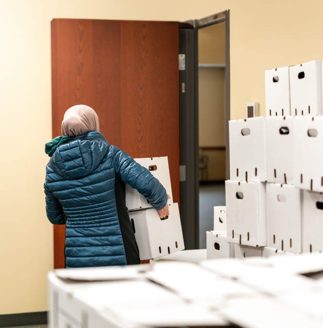 A woman in a blue coat and headscarf picks up two white boxes from a large stack of boxes