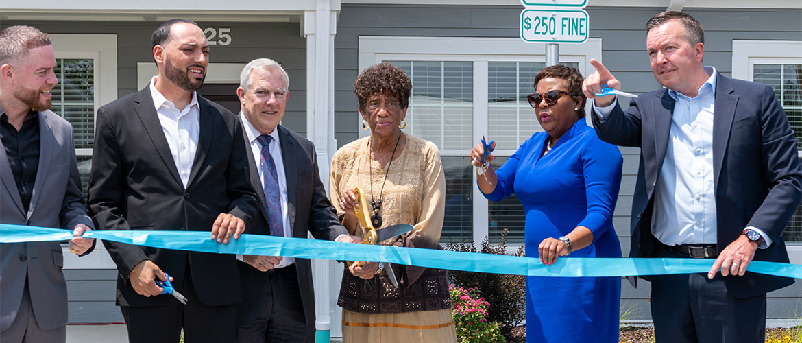 Four men and two women in business attire prepare to cut a ceremonial ribbon in front of a new housing development.