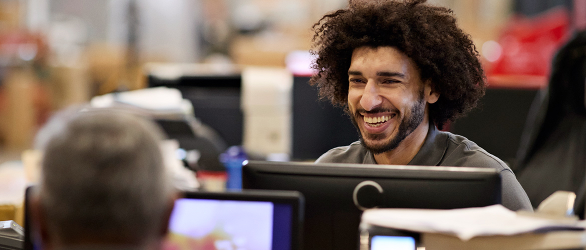 a women in a light blue suit sits at a desk smiling next to an older man in a suit