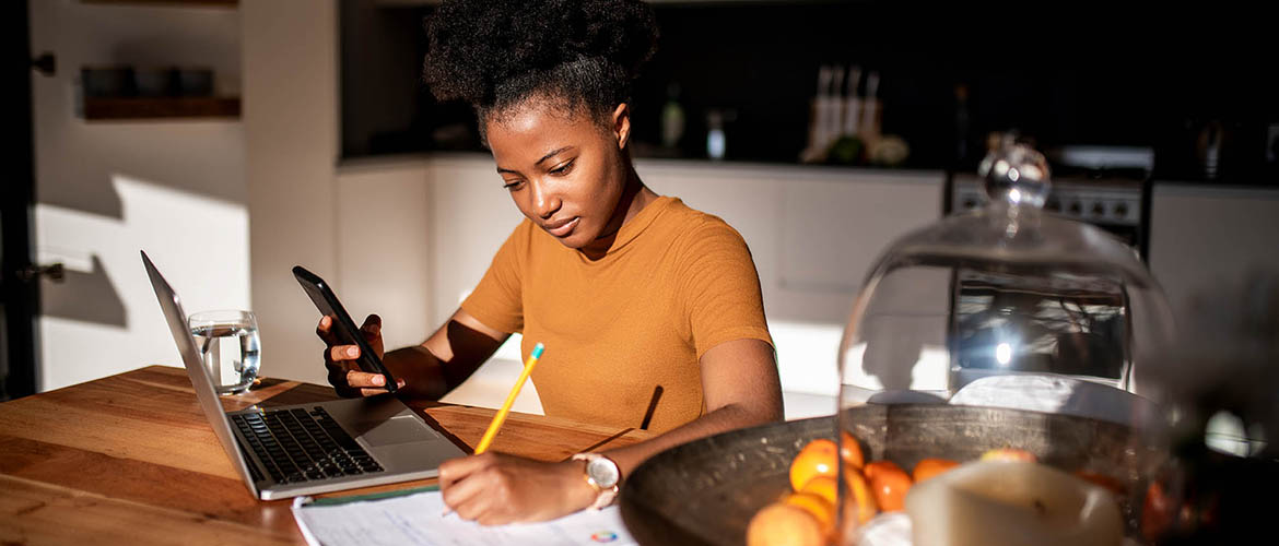 young woman in kitchen takes notes from phone and laptop computer 