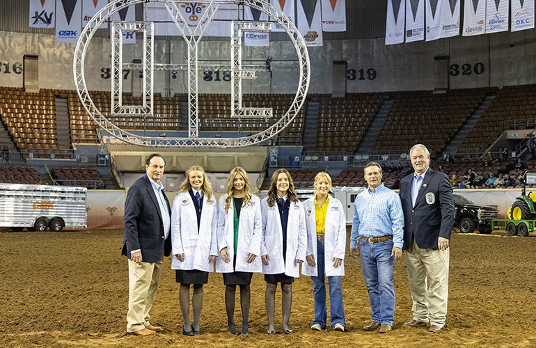 Four teen girls in white medical coats stand in an arena with a dirt floor for a livestock show