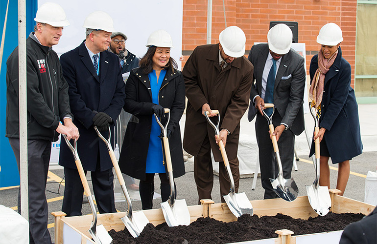 Men and women in coats and hard hats turn over dirt with shovels at a ceremonial groundbreaking. 